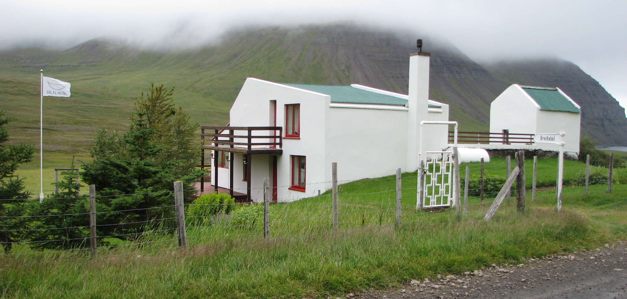 Breiðaból í Skálavík í júlí 2011. Öskubakur í baksýn, hulinn skýjum.  Áður var fleirbýlt á Breiðabóli en þetta hús er nú í eigu Karitasar Hafliðadóttur í Bolungarvík, ekkju Óskars Hálfdánarsonar og afkomenda þeirra. Húsið stendur líklega þar sem Neðribær stóð, sem var eitt Breiðabólsbýlið, en þar bjó Jón faðir Jónu, Ingimundar, Sigurðar og þeirra systkina síðustu tvo áratugina. Breiðaból kemur víða við þessa sögu.